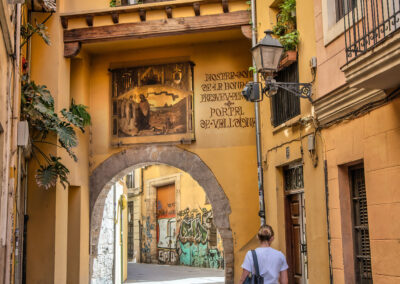 A woman in a white t-shirt walking through an archway of a yellow building with overlooking balcony