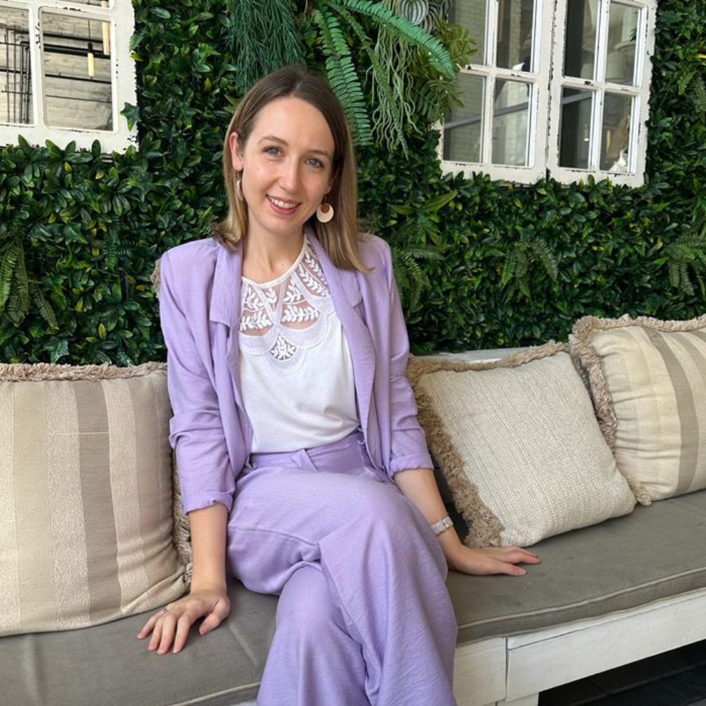 A woman in a purple trouser sit sat on a sofa next to green plants