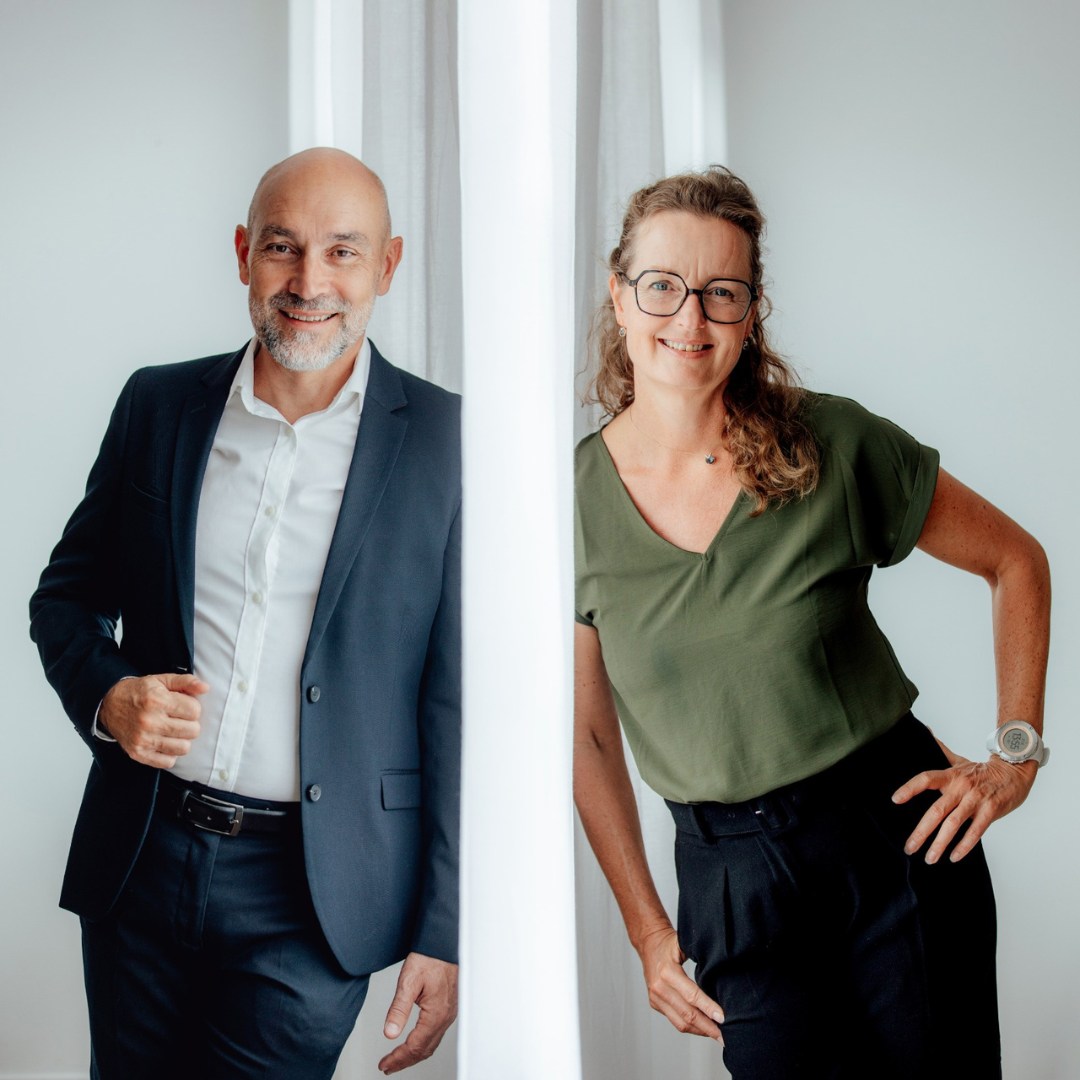 Two people stood posing for a photo on either side of a white wall