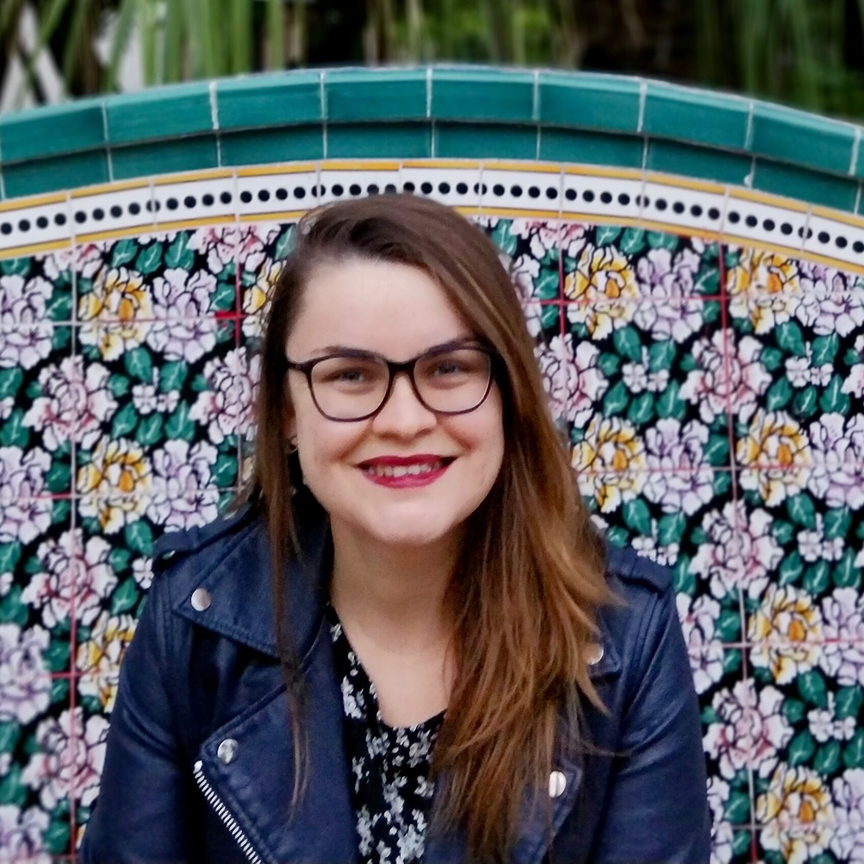 A woman with glasses smiling in front of colourful flower mosaic