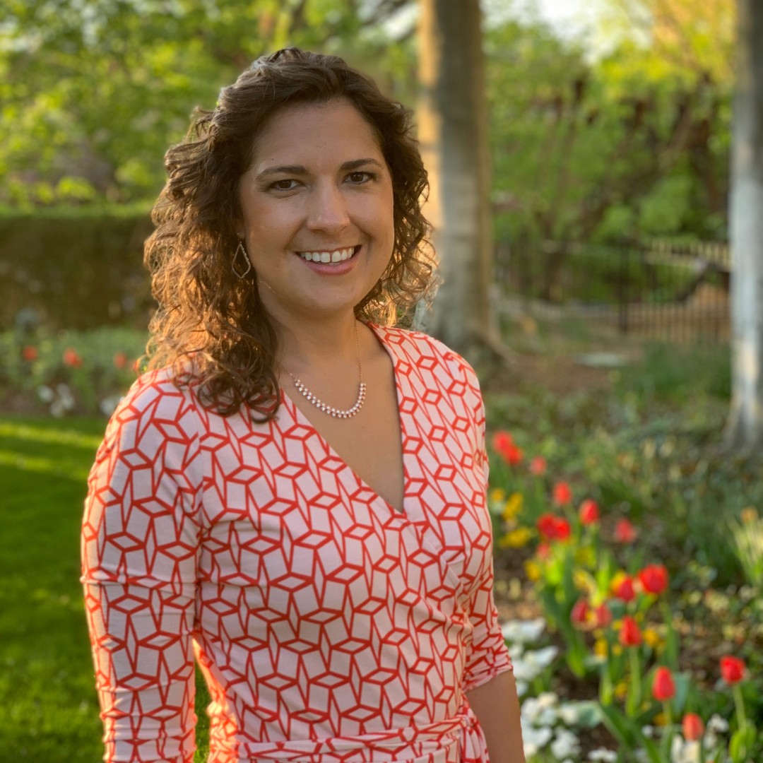 A young woman with curly brown hair wearing a red dress smiling in a garden