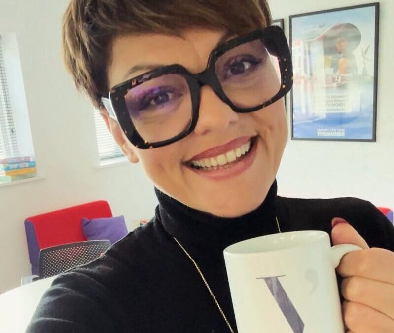 A woman in large square glasses taking a selfie holding a coffee
