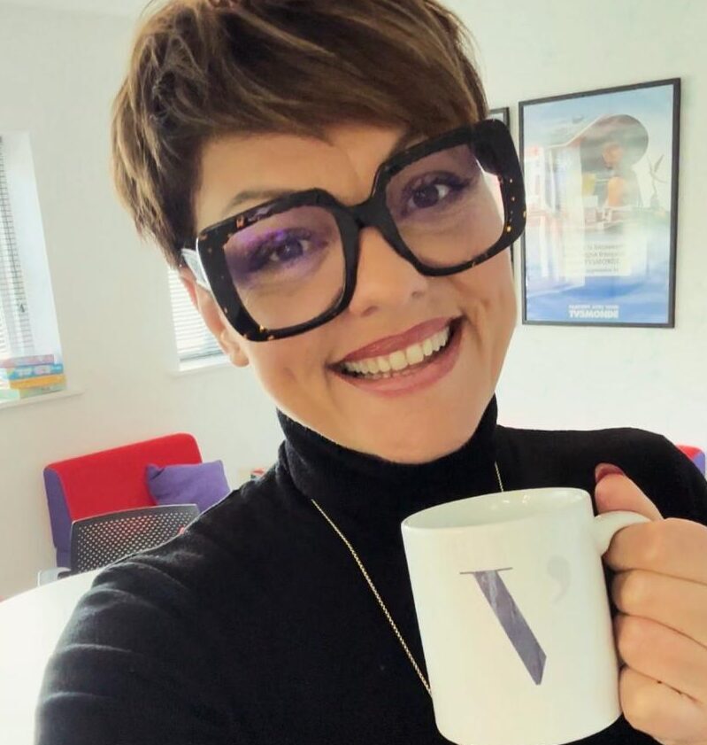 A woman in large square glasses taking a selfie holding a coffee